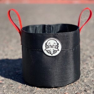 May include: A black fabric bucket with red handles and a white logo of a smiling face with a beard. The logo is on the front of the bucket.