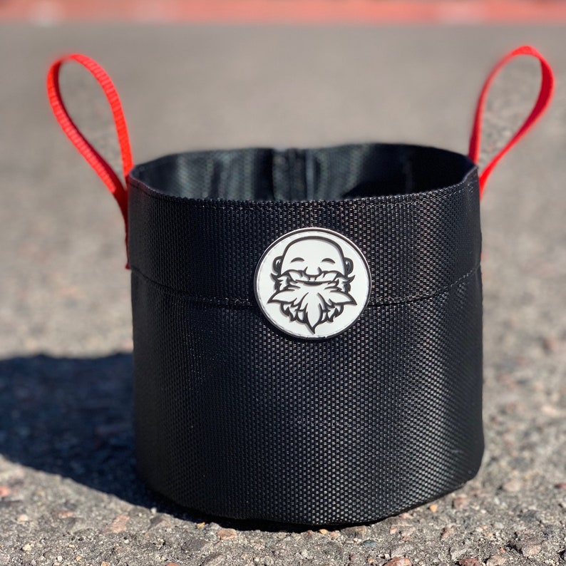 May include: A black fabric bucket with red handles and a white logo of a smiling face with a beard. The logo is on the front of the bucket.