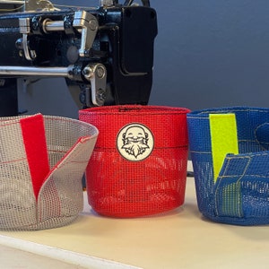 May include: Three mesh fabric containers in red, gray, and blue, each with a different colored Velcro closure. The red container has a circular emblem with a bearded face. A black sewing machine is in the background.