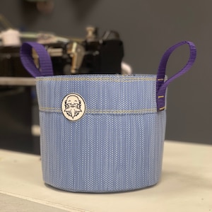 May include: A blue and white striped fabric planter with purple handles. The planter has a small white sticker with a cartoon face on it.