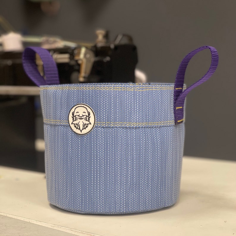 May include: A blue and white striped fabric planter with purple handles. The planter has a small white sticker with a cartoon face on it.