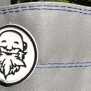May include: A white and black circular patch with a cartoon illustration of a person with a beard. The patch is attached to a gray fabric with blue stitching.