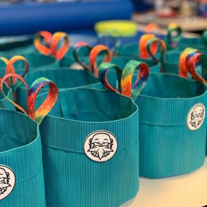 May include: A group of teal blue fabric storage containers with rainbow tie-dye handles. Each container has a white circle with a cartoon image of a bearded man with a smile.