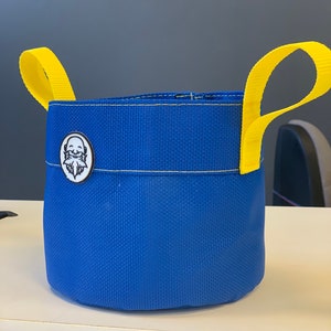May include: A blue canvas bucket with yellow handles and a white logo of a person with a beard. The bucket is made of a durable material and is perfect for carrying small items.