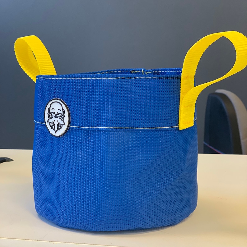 May include: A blue canvas bucket with yellow handles and a white logo of a person with a beard. The bucket is made of a durable material and is perfect for carrying small items.