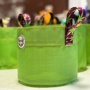 May include: Green mesh grow bag with a purple and green leaf print handle. The bag has a yellow stitched seam and a round sticker with a cartoon face on it.