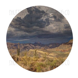 May include: A scenic view of a desert landscape with mountains in the distance. The sky is overcast with dark clouds. There are several tall cacti in the foreground.