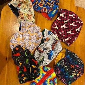 Op de afbeelding: Een verzameling van negen kleurrijke laskappen van stof met verschillende patronen, waaronder een rood-zwart ruitpatroon, een blauw-geel stripboekontwerp, een rood-wit-blauw tie-dye patroon, een zwart-groen peperpatroon, een kaart van Georgia, een zwart-wit autopatroon en een zwart-geel hertenpatroon.