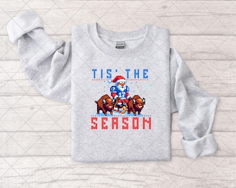 May include: Gray sweatshirt with a pixelated image of Santa Claus riding two buffaloes. The text "Tis' the Season" is printed in red.