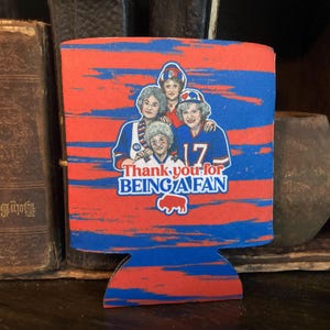 May include: A red and blue can cooler with a striped pattern. It features a group of four women wearing blue and red jerseys with the text "Thank you for being a fan" and a buffalo silhouette.