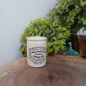 May include: A cylindrical off-white ceramic jar with a crackled finish. The front features black printed text reading: GRAND MEDAL OF MERIT VIENNA, JAMES KEILLER & SONS, DUNDEE MARMALADE, ONLY PRIZE MEDAL FOR MARMALADE, LONDON 1862, with decorative flourishes.