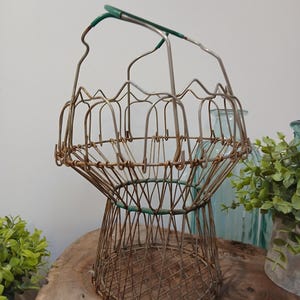 May include: A vintage wire egg basket with a handle. The basket is made of metal wire with a brown, aged finish. The handle and some accents are painted green. The basket has a unique shape, with a wide base and a narrower top.