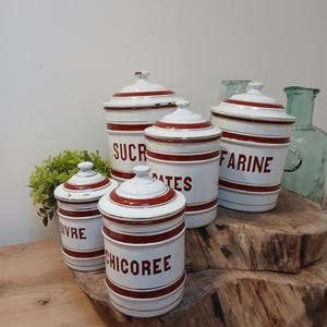 Pode incluir: Um conjunto de potes de cerâmica branca com listras e tampas vermelhas. Os potes são rotulados com palavras francesas como "SUCRE" e "FARINE". Eles são dispostos em uma superfície de madeira, sugerindo uma estética vintage ou rústica.