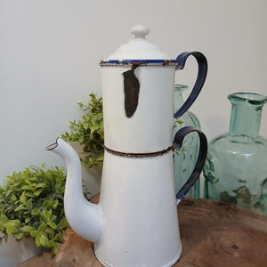 May include: White enamel coffee pot with a blue rim and handle. The vintage pot has a spout and a lid with a small knob. The pot is sitting on a wooden surface with green plants and glass bottles in the background.
