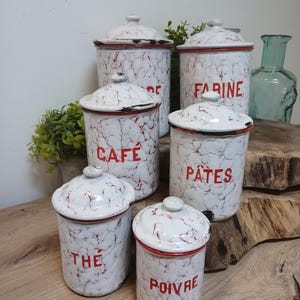 May include: A set of white enamel kitchen storage jars with red marbling and red trim. The jars are labelled with the words "CAFE", "FARINE", "PATES", "THE", and "POIVRE" in red lettering. The lids have small knobs.