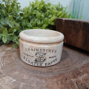 May include: A cylindrical ceramic pot with a cream-colored crackle glaze finish. The front features black printed text reading J. SAINSBURY'S FRESHLY MADE BLOATER PASTE with a central S logo containing a small floral illustration and trade mark text.