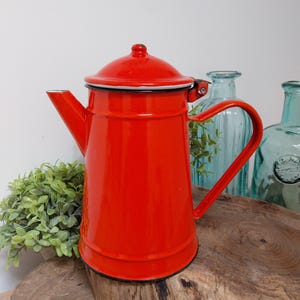 Puede incluir: Una cafetera de esmalte rojo brillante con ribete negro, con tapa y asa curvada. La cafetera se exhibe sobre una superficie de madera rústica, con follaje verde y botellas de vidrio azul al fondo. Un artículo de cocina clásico.