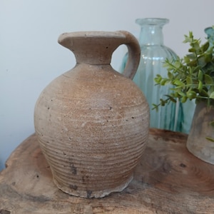 May include: A beige ceramic pitcher with a handle and spout. The pitcher has a rounded body with horizontal ridges and a textured surface. It is displayed on a wooden surface, suggesting a vintage or farmhouse aesthetic. The pitcher is likely made of earthenware.