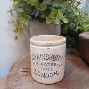 Puede incluir: Un tarro cilíndrico de cerámica color hueso con un borde superior. La parte frontal presenta un texto impreso en negro que dice J. SAINSBURY BLOATER PASTE LONDON. La superficie del tarro muestra signos de antigüedad con manchas oscuras.