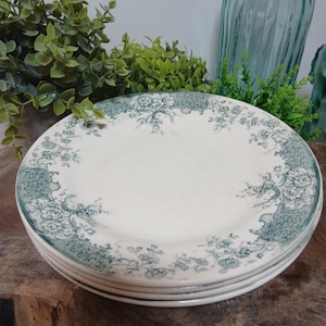 May include: A stack of round ceramic plates featuring an off-white base color and a decorative border pattern in dark green. The intricate design consists of floral motifs, scrolling vines, and textured detailing along the outer rim of each plate.