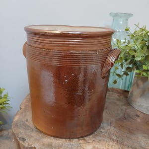 Może przedstawiać: Brązowy ceramiczny garnek z błyszczącym wykończeniem. Garnek ma szeroki, żebrowany brzeg i dwa małe uchwyty. Stoi na drewnianej powierzchni. Jasnoniebieski szklany wazon i zieleń w tle.