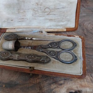 May include: A vintage sewing kit presented in a brown leather case. The kit contains silver-toned scissors, a thimble, a needle, and a small case. The tools feature intricate floral patterns. The interior of the case is lined with cream-colored fabric.