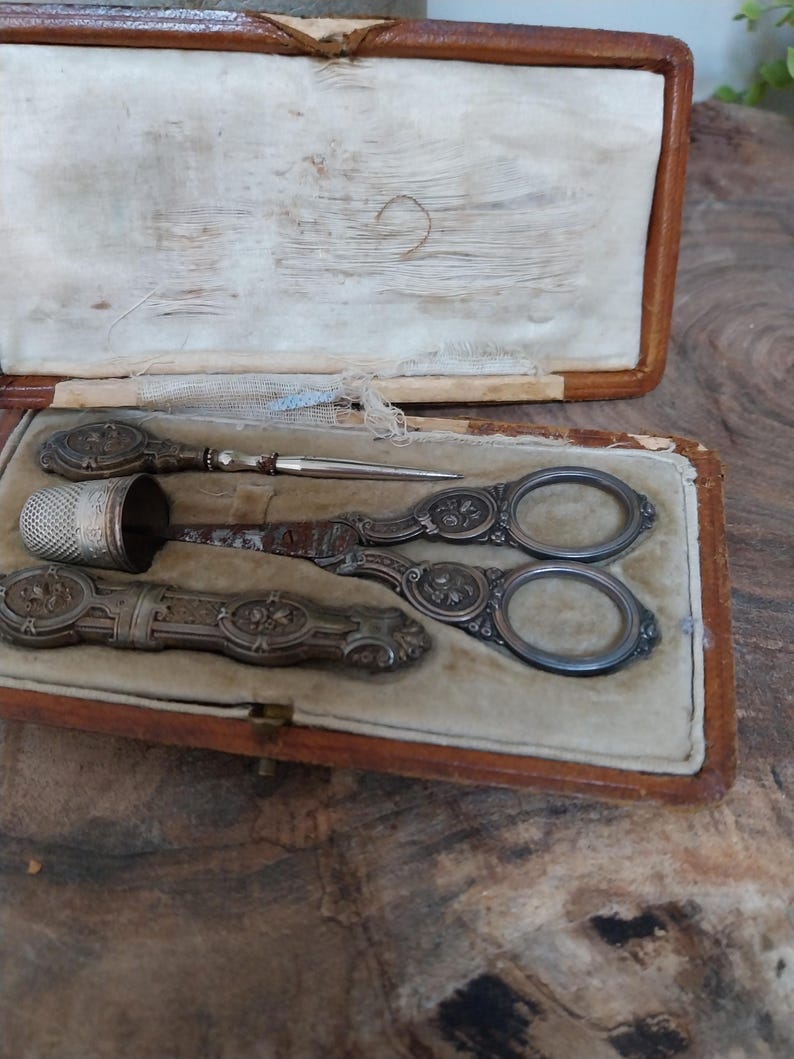 May include: A vintage sewing kit presented in a brown leather case. The kit contains silver-toned scissors, a thimble, a needle, and a small case. The tools feature intricate floral patterns. The interior of the case is lined with cream-colored fabric.