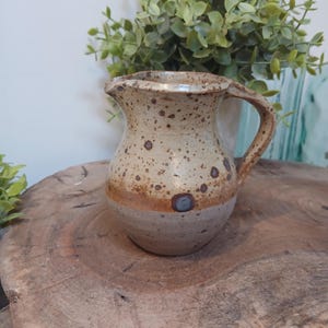 May include: A small, speckled ceramic pitcher with a handle. The pitcher has a light tan base with brown speckles and a darker brown band around the middle. It sits on a wooden surface, with greenery in the background.