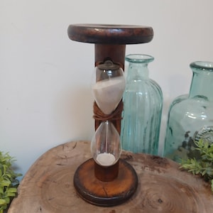 May include: A vintage hourglass with a wooden frame and base. The hourglass has two clear glass bulbs connected by a narrow neck, filled with white sand. The wooden frame is a dark brown colour. Decorative glass bottles are in the background.