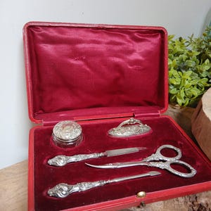 Könnte beinhalten: Ein Vintage-Toilettenset in einem rechteckigen, mit rotem Samt ausgekleideten Etui. Das Set enthält ein kleines Glasgefäß mit Silberdeckel, einen Brieföffner mit Silbergriff, eine verzierte Silberschere, einen Nagelhautschieber und einen Silberdeckel.