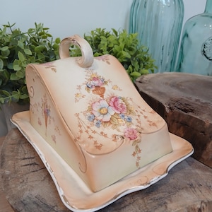 May include: A cream-colored ceramic butter dish with a domed lid and a rectangular base. The lid features a painted floral design with pink, blue, and yellow flowers, accented by gold trim along the edges and a curved handle on top.