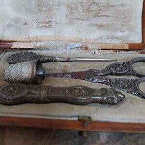 May include: An antique sewing kit in a wooden box with a beige velvet lining. The kit contains a thimble, scissors, a needle, and a long, ornate tool. The tools are silver-toned with floral designs. The box is closed with a small brass clasp.