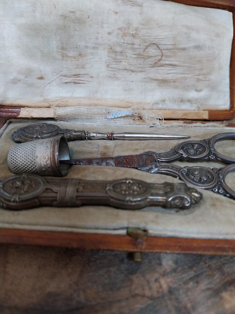 May include: An antique sewing kit in a wooden box with a beige velvet lining. The kit contains a thimble, scissors, a needle, and a long, ornate tool. The tools are silver-toned with floral designs. The box is closed with a small brass clasp.