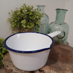 May include: White enamel saucepan with a blue rim and handle. The saucepan is set on a wooden surface, with two light blue glass bottles and greenery in the background. A vintage-style kitchenware item.