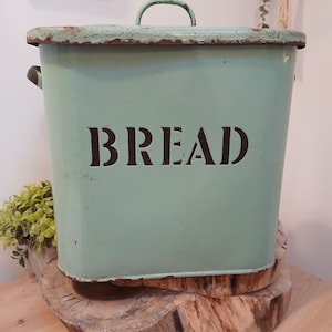 Puede incluir: Una panera rectangular de estilo vintage en un tono verde suave, con la palabra "BREAD" en letras oscuras y en negrita. La panera tiene una tapa con asa y asas laterales. Los bordes muestran signos de desgaste, lo que le da un aspecto rústico. Se encuentra sobre una base de madera.