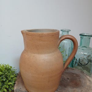 May include: A tan-colored ceramic pitcher with a handle and spout. The pitcher has a textured surface and decorative stitched details around the top and middle. It sits on a wooden surface, with green and blue glass bottles in the background.
