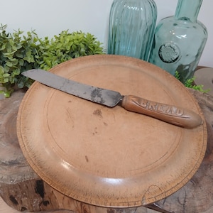 May include: A vintage bread knife with a weathered metal blade and a wooden handle featuring the word BREAD embossed into the side. The knife rests horizontally across a round, light-coloured wooden plate with visible surface wear and grain.