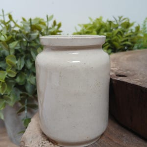 May include: A cylindrical ceramic jar with a smooth, off-white glaze and subtle speckled texture. The vessel features a slightly flared rim and a straight body, presenting a neutral appearance with a consistent finish across its exterior surface.