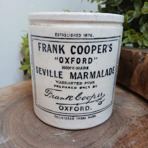Op de afbeelding: Een cilindervormige witte keramische pot met zwarte bedrukte tekst die Frank Cooper's Oxford Seville Marmalade, Established 1874, Warranted Pure, Prepared Only By Frank Cooper, Oxford, Registered Trade Mark leest, met een gespikkelde afwerking.