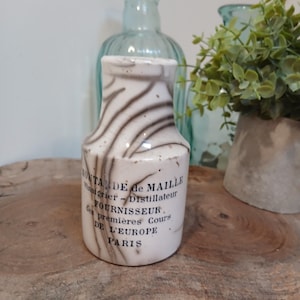 Puede incluir: Una pequeña botella de cerámica blanca con un patrón marrón arremolinado único. La botella tiene forma cilíndrica con cuello estrecho y parte superior acampanada. Se ve texto negro en la botella, lo que indica que es un producto de París.