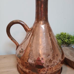 May include: A large, hammered copper pitcher with a wide, rounded base that tapers into a tall, narrow neck. The vessel features a single curved handle attached to the side and a flat, circular base. The surface has a reflective, metallic finish.