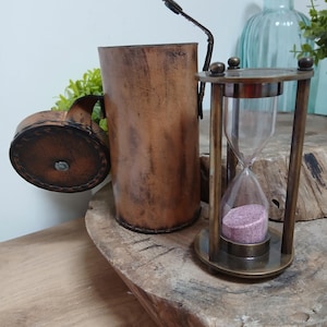 Puede incluir: Un reloj de arena de estilo vintage con marco de latón y arena rosa, junto a un recipiente de cuero marrón y un jarrón decorativo. Un objeto de decoración clásico y atemporal.