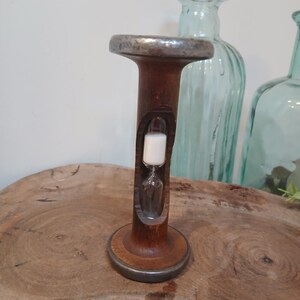 Può includere: Una clessidra vintage con cornice in legno e dettagli in metallo. La clessidra contiene sabbia bianca ed è appoggiata su una superficie di legno. La clessidra è alta circa 15 cm.