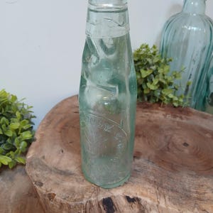 Puede incluir: Una botella de vidrio verde claro vintage con un diseño texturizado único. La botella tiene una forma redondeada y un cuello estrecho. La botella tiene una etiqueta en relieve. La botella se exhibe sobre una superficie de madera.