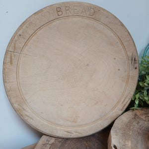 Peut inclure: Une planche à pain ronde en bois clair avec le mot BREAD gravé sur le bord supérieur. La surface présente un grain de bois naturel, des marques de couteau visibles et un détail de rainure circulaire le long du centre plat de la planche.