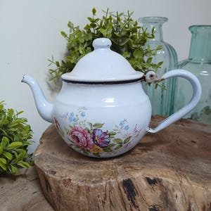 Könnte beinhalten: Weiße Emaille-Teekanne mit einem Blumendesign mit rosa, lila und blauen Blumen. Die Teekanne hat einen gebogenen Ausguss, einen Griff und einen Deckel mit einem kleinen Knopf. Der Rand und die Griffbefestigungen sind dunkelbraun.