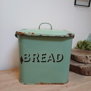 Może przedstawiać: Prostokątny, miętowy pojemnik na chleb z otwieraną pokrywą i uchwytem. Słowo "BREAD" jest wycięte z przodu. Metalowy pojemnik na chleb ma postarzany, rustykalny wygląd z widoczną rdzą.