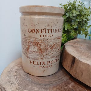 May include: A cylindrical beige ceramic jar with a brown printed label featuring the text CONFITURES FINES FELIX POTIN PARIS and an illustration of a factory complex. The jar has a smooth finish with subtle speckling and a recessed top rim.