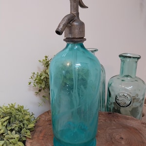 Op de afbeelding: Een vintage turquoise glazen selterfles met een metalen dispenser. De fles is cilindrisch met een afgeronde basis en een lange hals. De dispenser heeft een hendel en een tuit. Andere glazen flessen staan op de achtergrond.