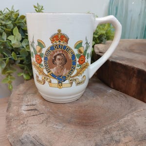 Op de afbeelding: Een witte keramische mok met een centraal portret van koningin Elizabeth II, omringd door het koninklijk wapen, inclusief een kroon, het motto van de Kousenbandorde Honi Soit Qui Mal Y Pense en de tekst Dieu Et Mon Droit met bloemmotieven.